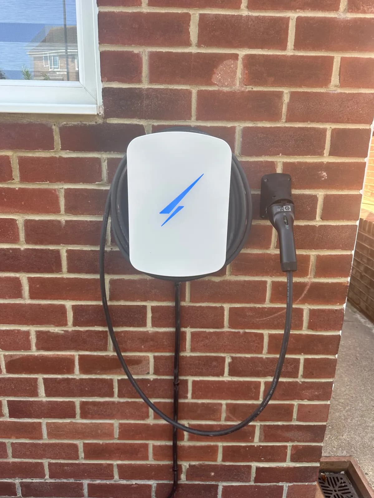 White tethered EV charger with a blue light, mounted on a red brick wall under a window